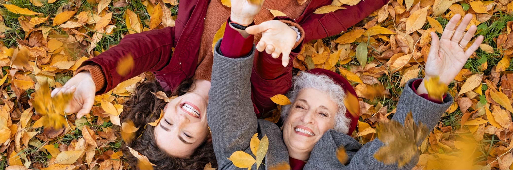Fall Into Fall Email Banners-3 Two smiling women lying in a bed of fall leaves, with their hands up in celebration.