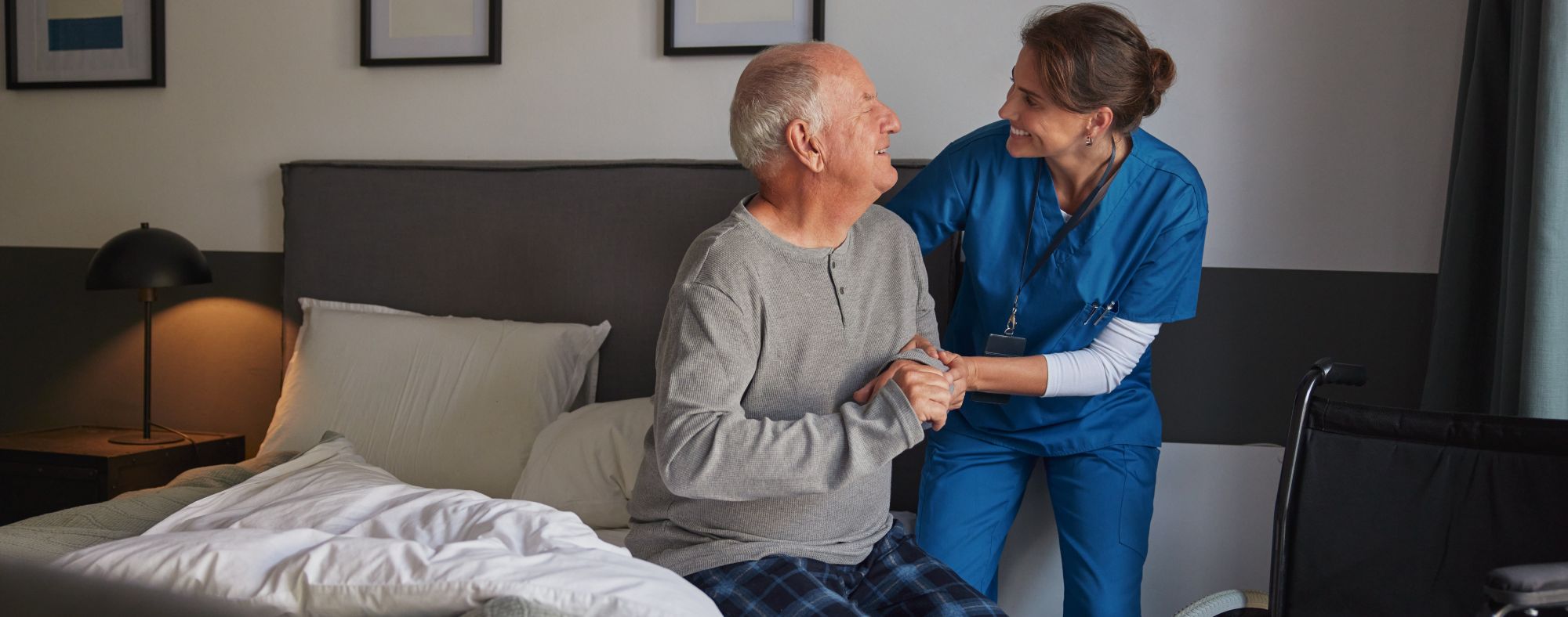 AdobeStock_1375444088-crop A senior man is sitting in bed and smiling at a caregiver, who is smiling back and helping him up.