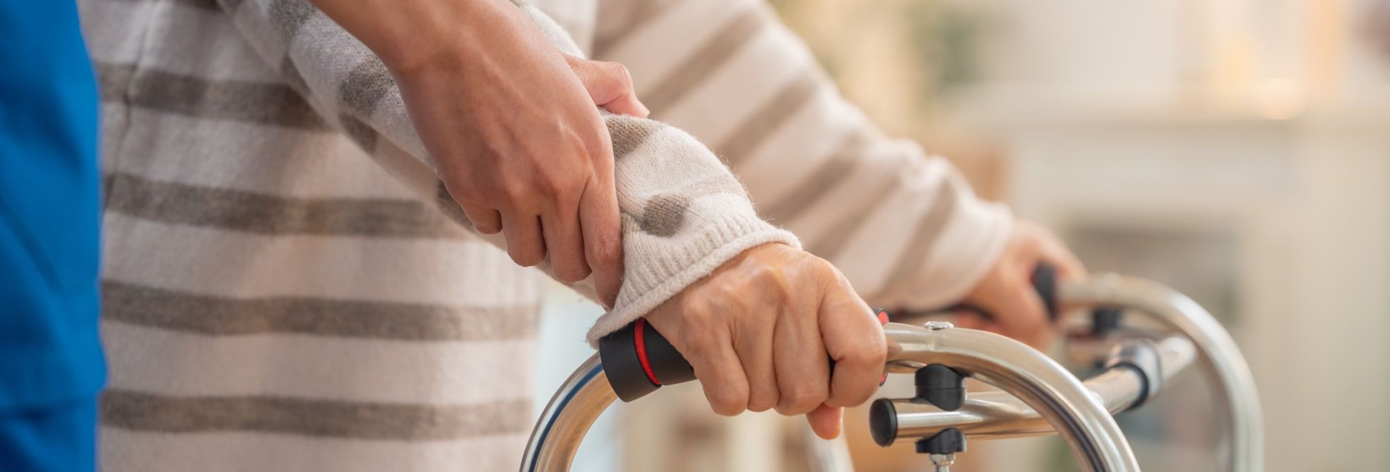 shutterstock_2653638571-crop A caregiver holds and guides a senior's wrist as the senior uses a walker.