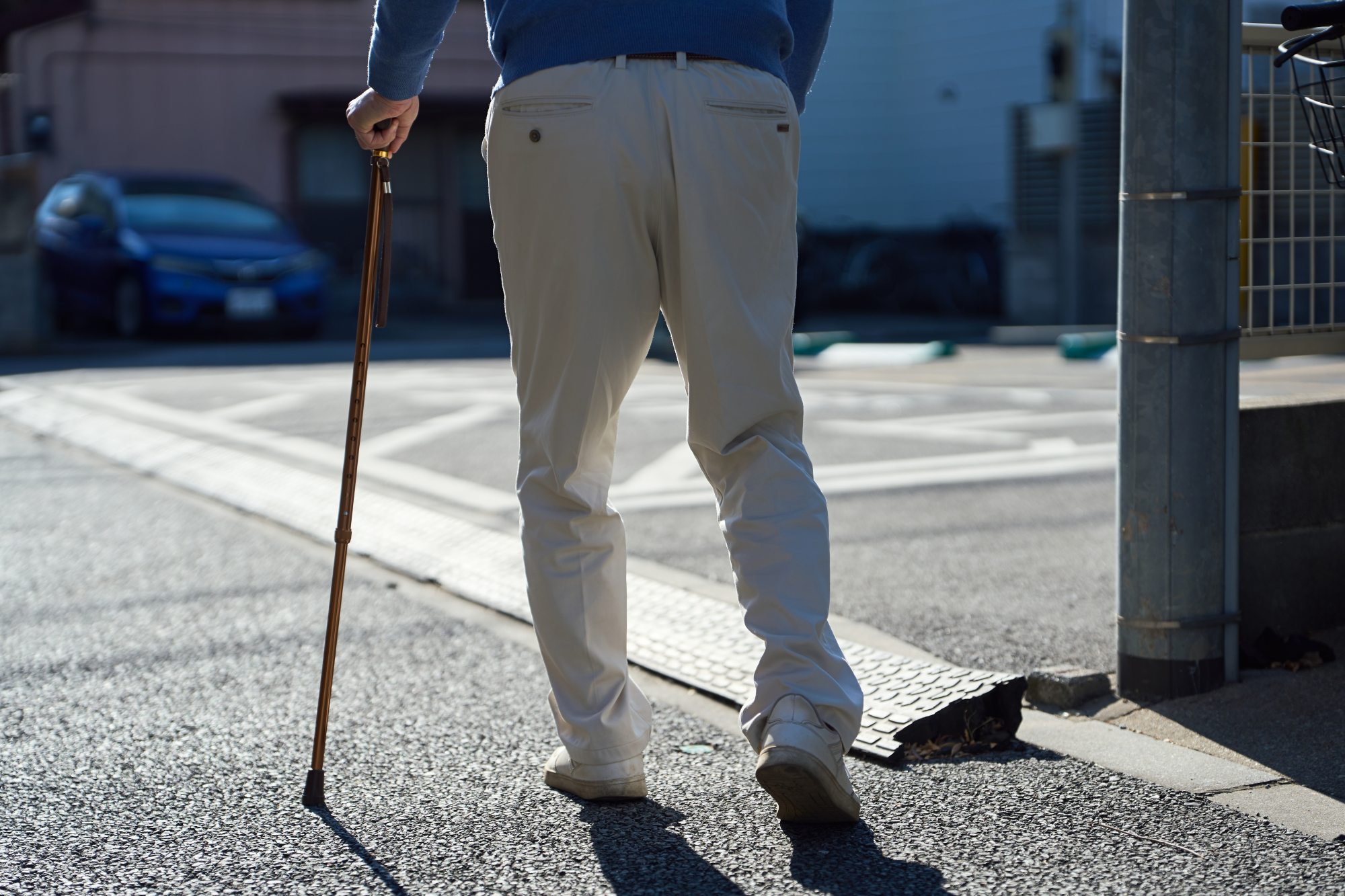 An older man is out on the street. He is shown from the back, waist down, with one foot walking forward. He holds a cane and his footsteps seem uncertain.